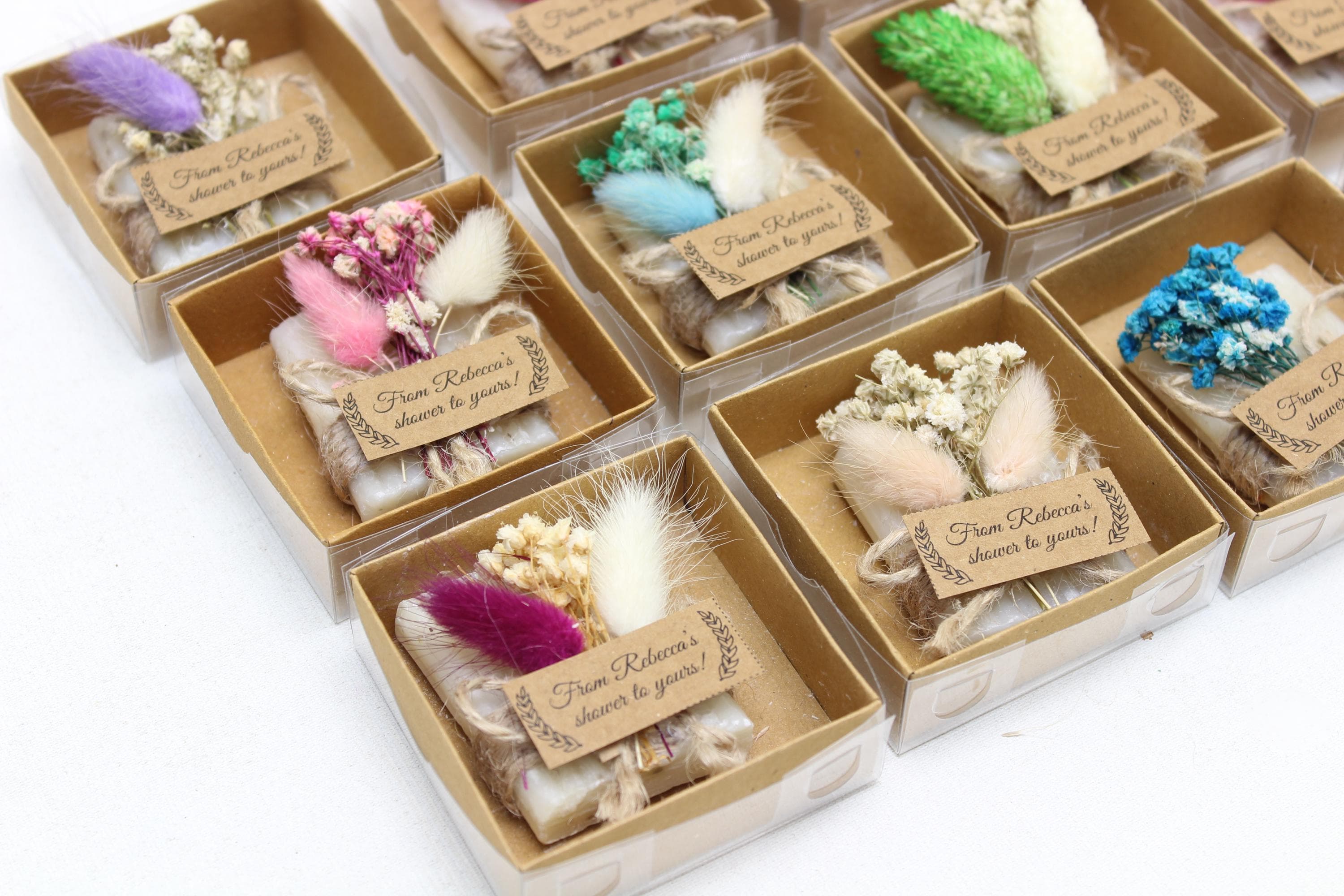 Square gift boxes with clear lids holding soap bars wrapped in twine with dried flowers and a personalised shower tag