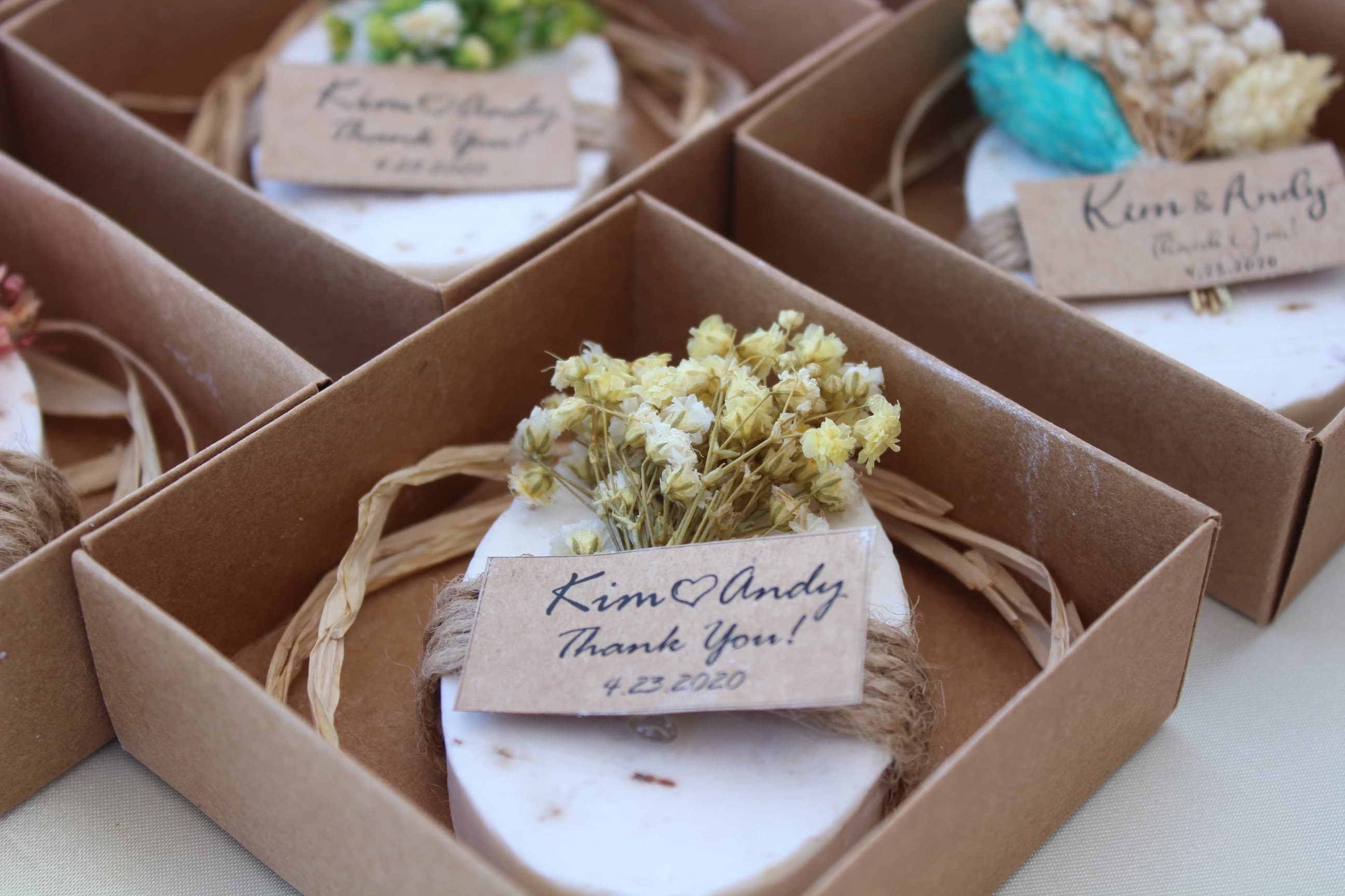 White oval soap favours with dried flowers in kraft boxes, each tied with twine and a personalised thank-you tag with names and wedding date
